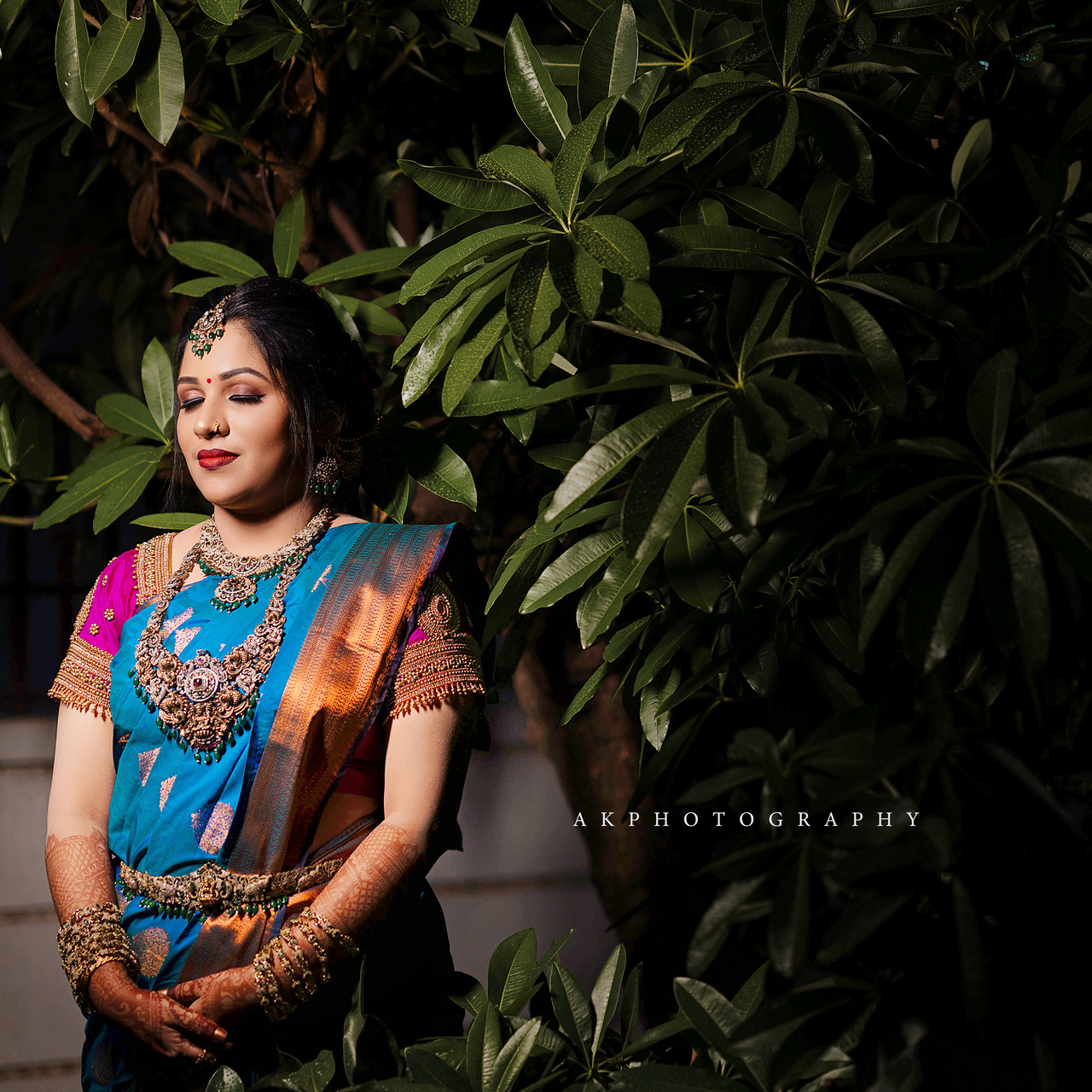 Young Indian woman in a traditional South Indian bridal saree with teal blue and gold embroidery, magenta blouse, and emerald and diamond jewelry, set against a lush green backdrop. 